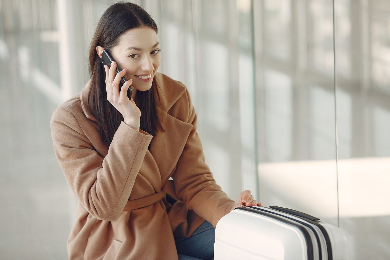 Young dreamy brunette in beige coat with suitcase having conversation via mobile phone while waiting for flight in airport terminal
